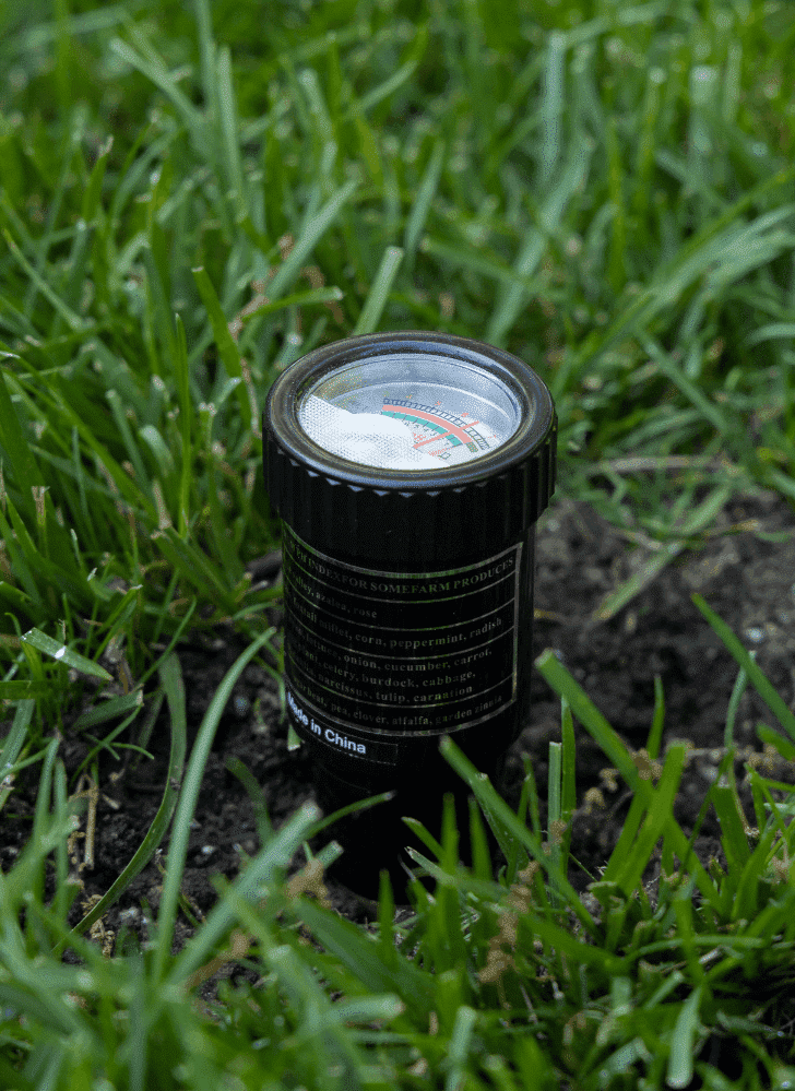 A Pro pH & Moisture Soil Tester embedded in the soil among grass, showing a close-up of its face and detailed markings.