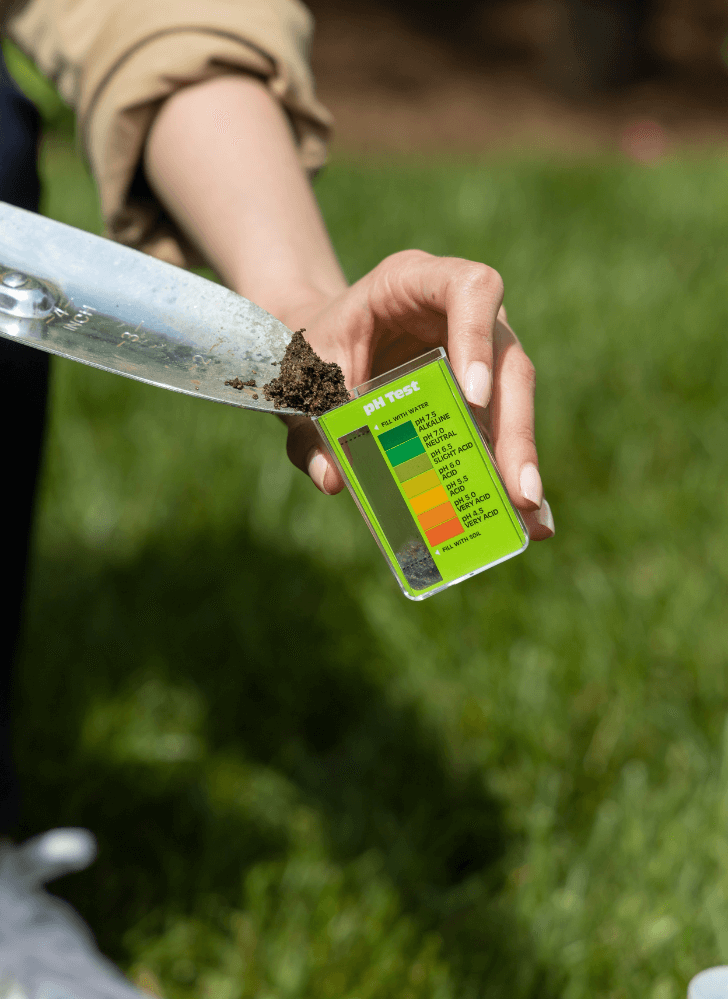 A person outdoors testing the pH level of soil using a Soil pH Test Kit for Lawns, specifically focusing on a scoop of dirt being analyzed.