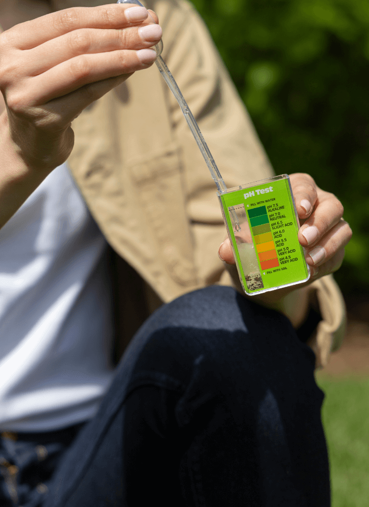 A person sitting on grass, using a Soil pH Test Kit for Lawns and comparing the results against a color chart to check the soil acidity.