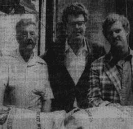 Three men standing close together, dressed in casual clothing, with blurred details in the background. The image appears to be in black and white.