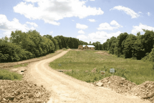 Gravel road leading to a distant house surrounded by trees under a partly cloudy sky.