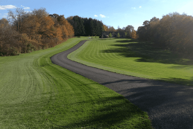 A winding asphalt path through a lush green park with autumn trees in the background under a clear sky.