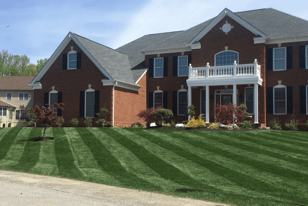 A large brick house with a white balcony and dark shutters showcases the best lawn mowing patterns, boasting a well-maintained lawn with diagonal stripes.