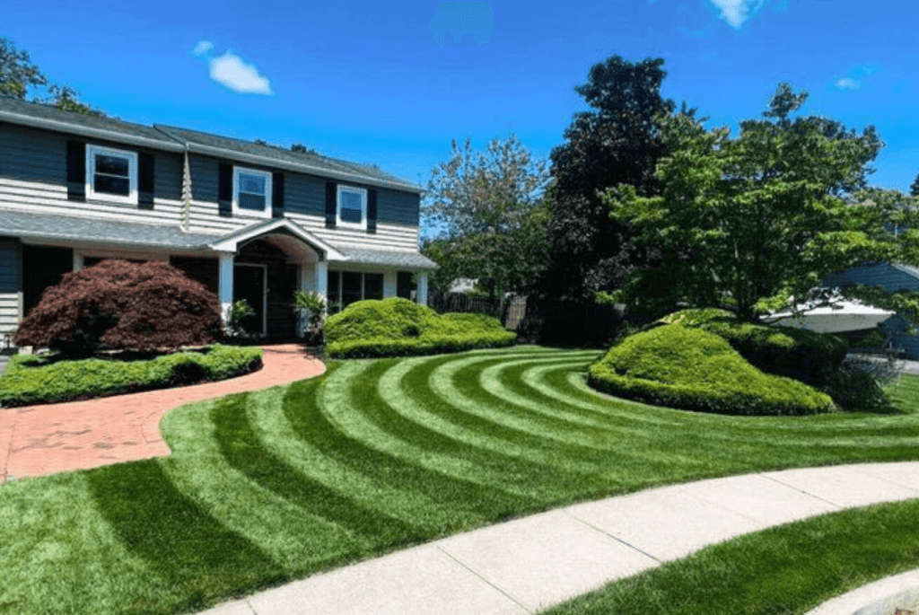 A suburban house with a neatly mowed lawn showcasing the best lawn mowing patterns, red brick pathway, and landscaped bushes under a clear blue sky.