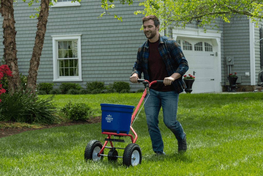 A man in a plaid shirt uses a lawn spreader on a grassy lawn in front of a gray house with white trim.