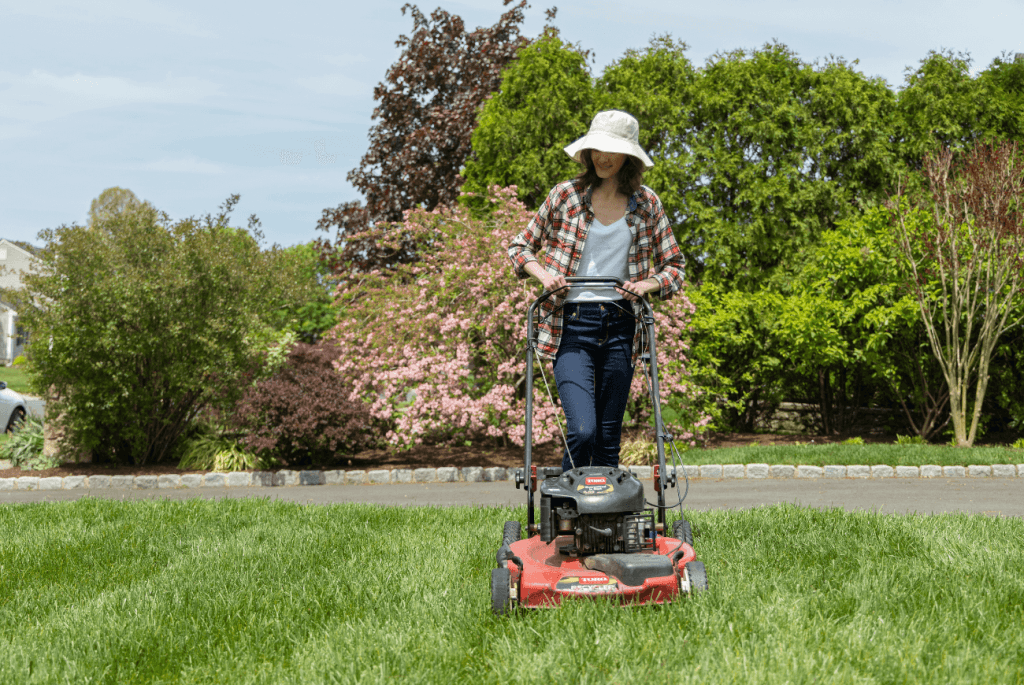 Wearing a sun hat and plaid shirt, the person is diligently preparing your lawn for winter as they mow the grass with a red lawnmower in the garden.