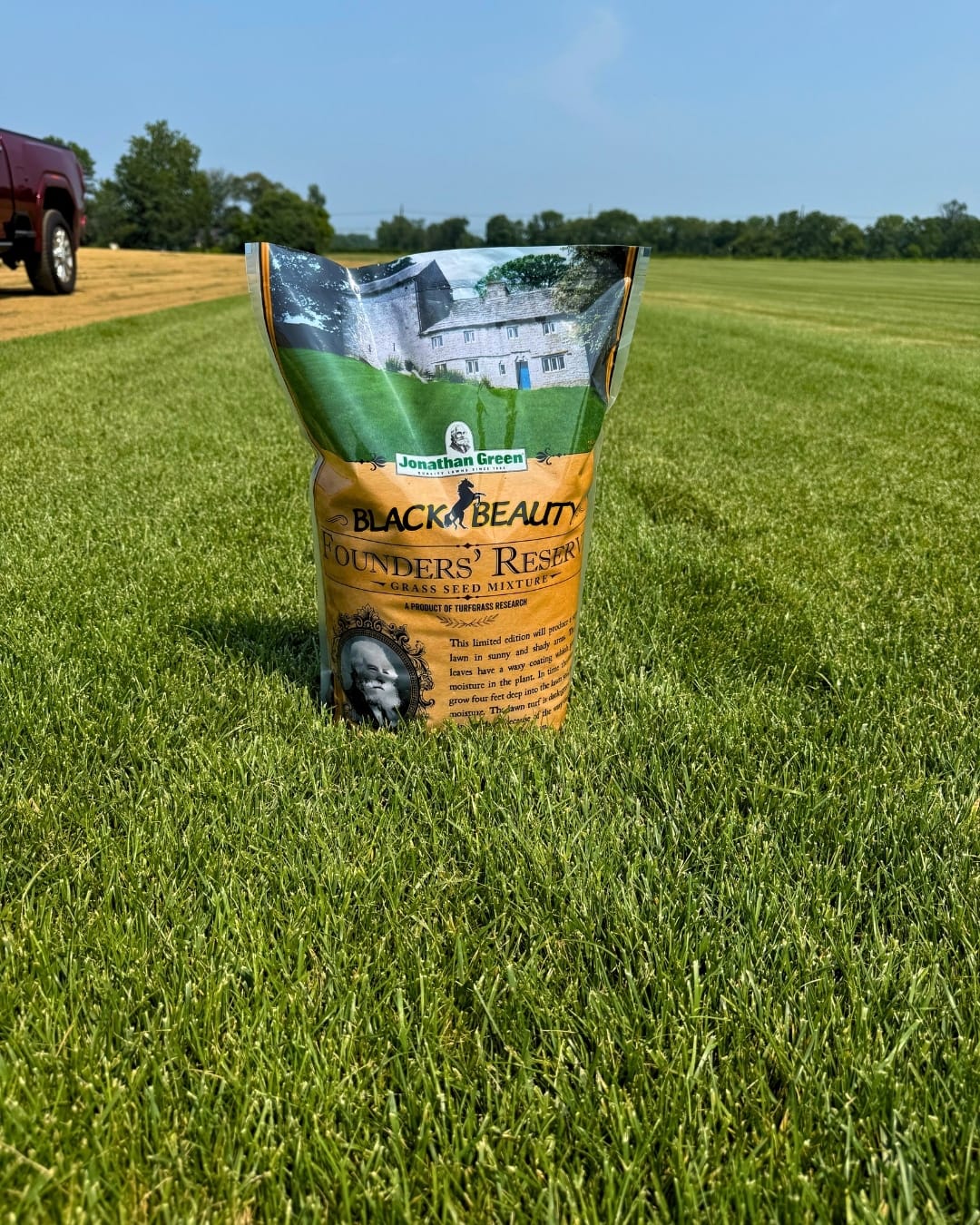 A bag of Black Beauty® Founders Reserve Grass Seed sits on a manicured lawn, with a truck and trees in the background.