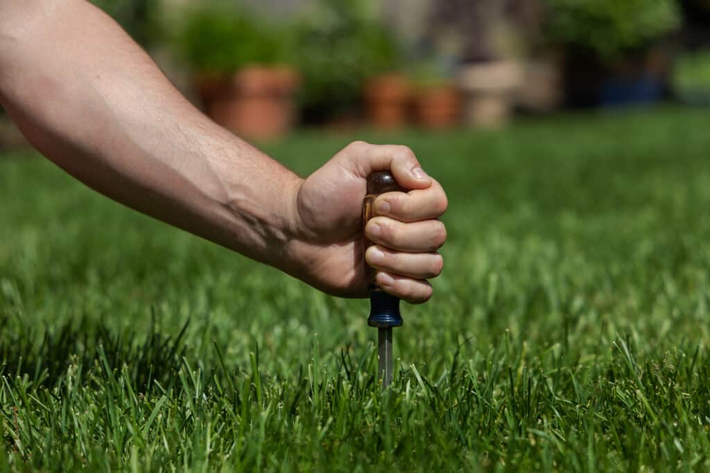 A person presses a soil probe or screwdriver vertically into green grass, with potted plants blurred in the background.